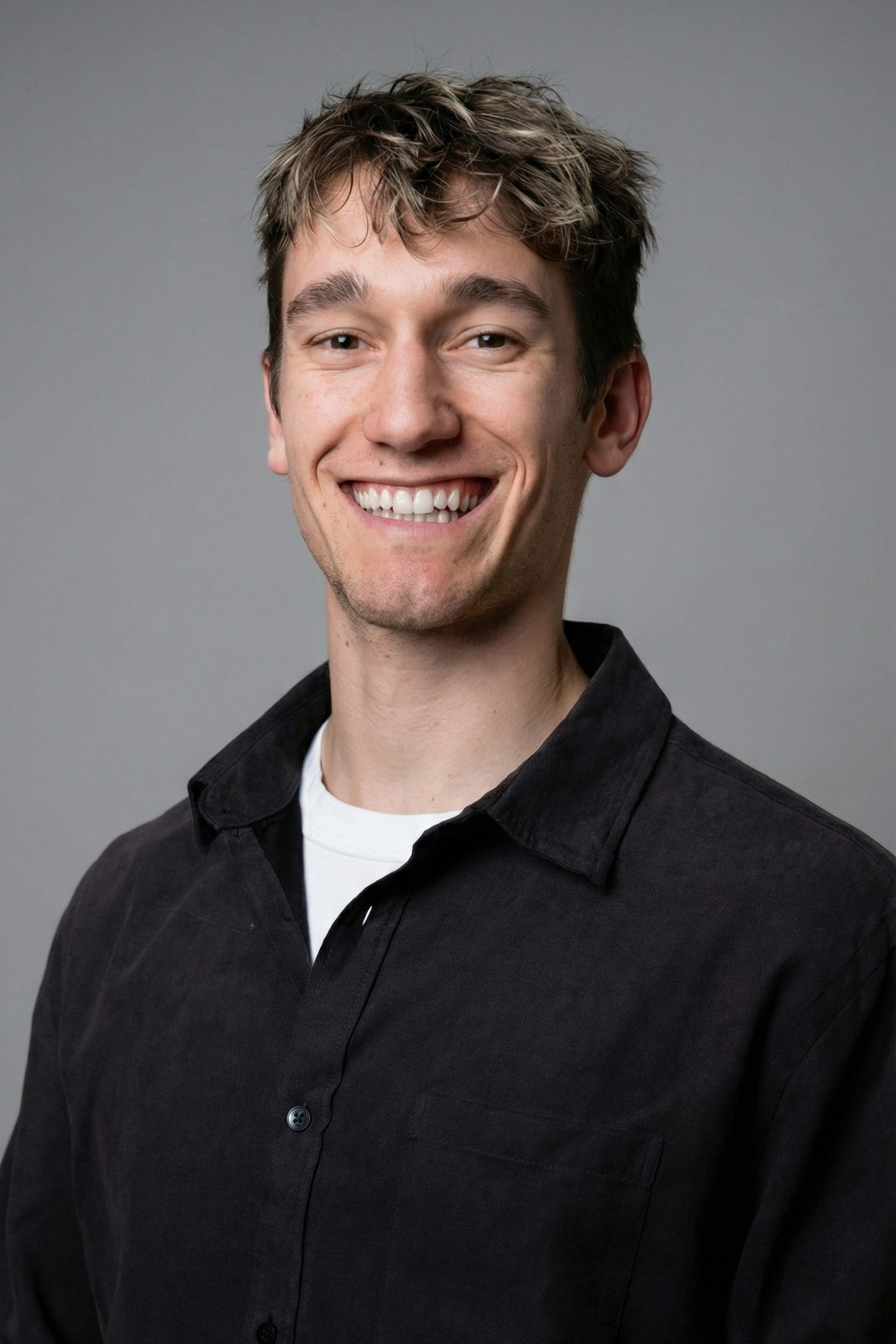 Young man with light hair wearing a white t-shirt and black watch, standing with arms crossed against a plain background.