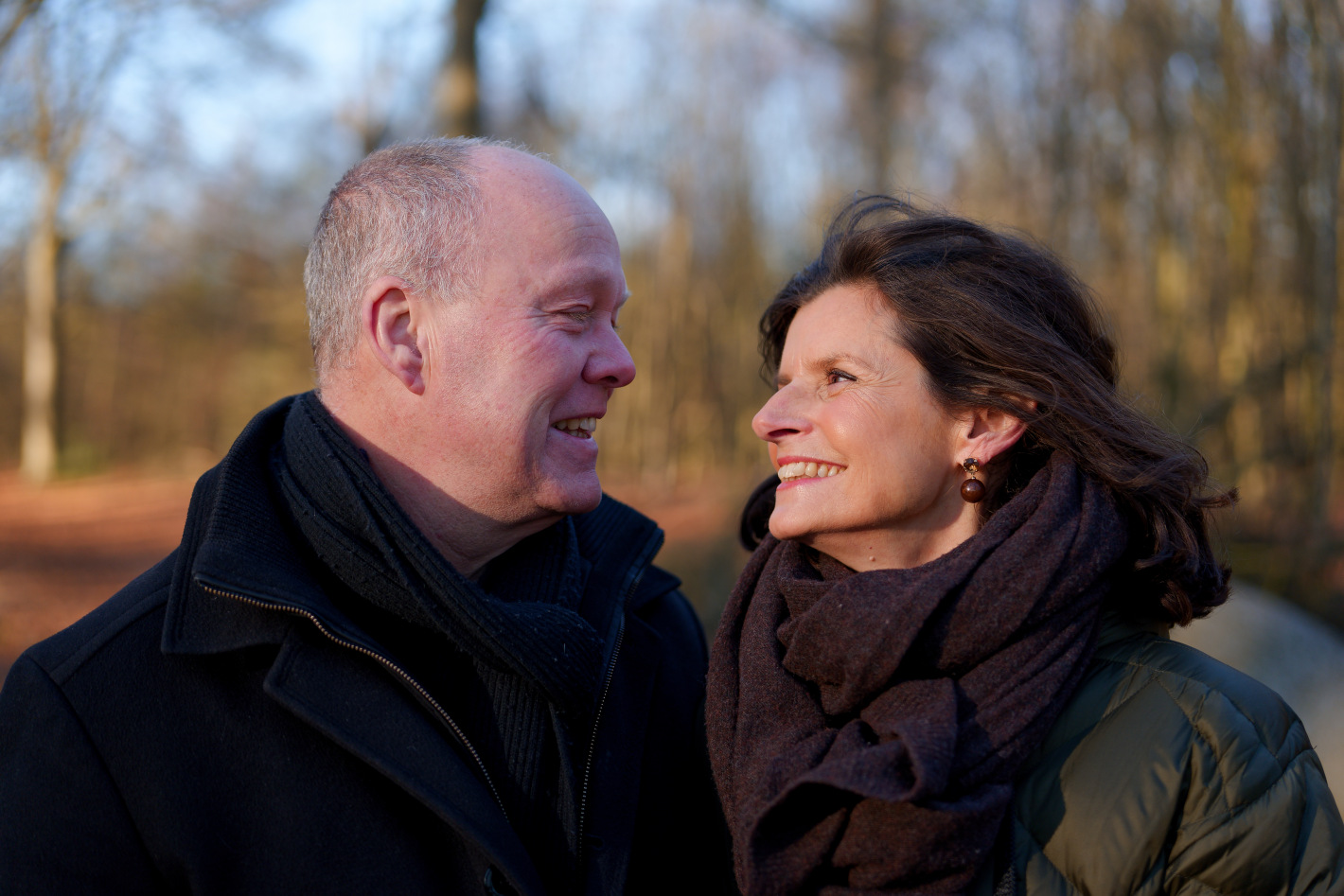 Oudere man en vrouw lachen naar elkaar buiten in een herfstachtig bos.