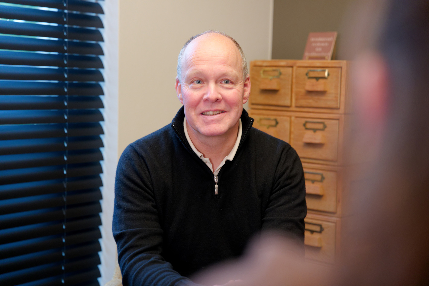 Man met een zwarte trui glimlacht terwijl hij zit in een kamer met houten ladenkast en jaloezieën.