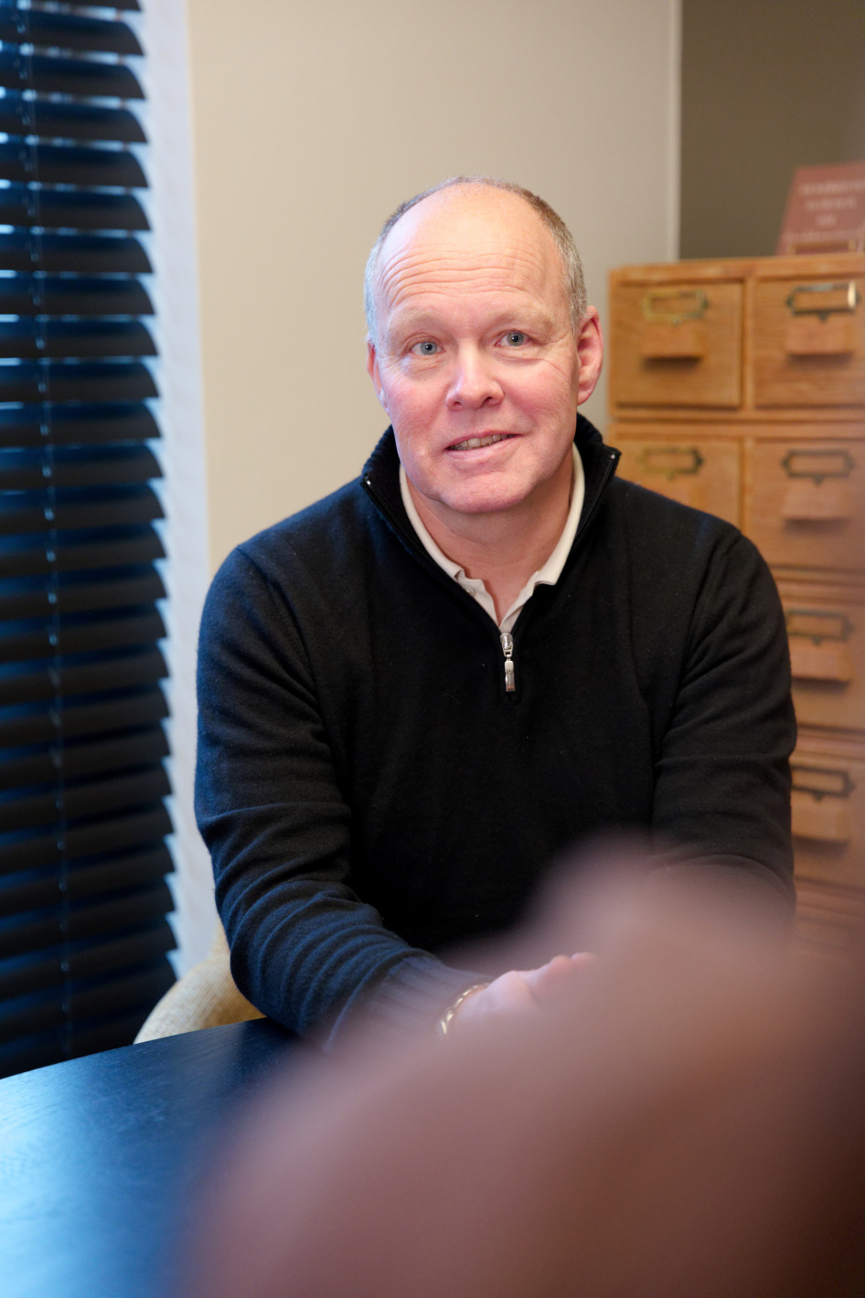 Man in een zwarte trui kijkt aandachtig terwijl hij aan een tafel zit in een kamer met houten ladenkast en donkere jaloezieën.