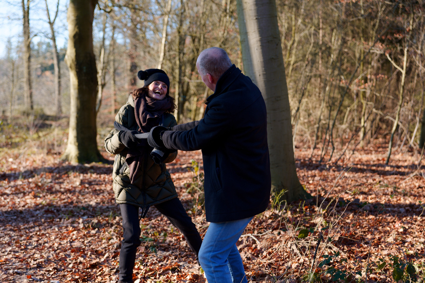 Twee volwassenen in winterkleding lachen en houden elkaars handen vast in een bos met gevallen bladeren.