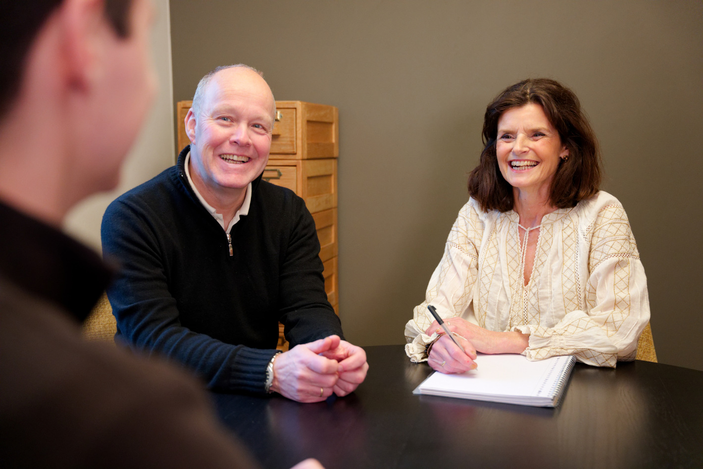 Twee glimlachende volwassenen, een vrouw schrijft met een pen in een notitieboek terwijl ze een gesprek hebben met een derde persoon.