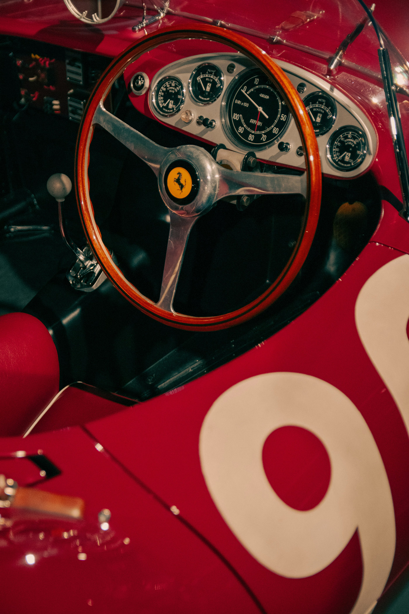 Close-up of a vintage red Ferrari car interior showing a wooden steering wheel with the Ferrari logo, dashboard gauges, and a white gear shift knob.