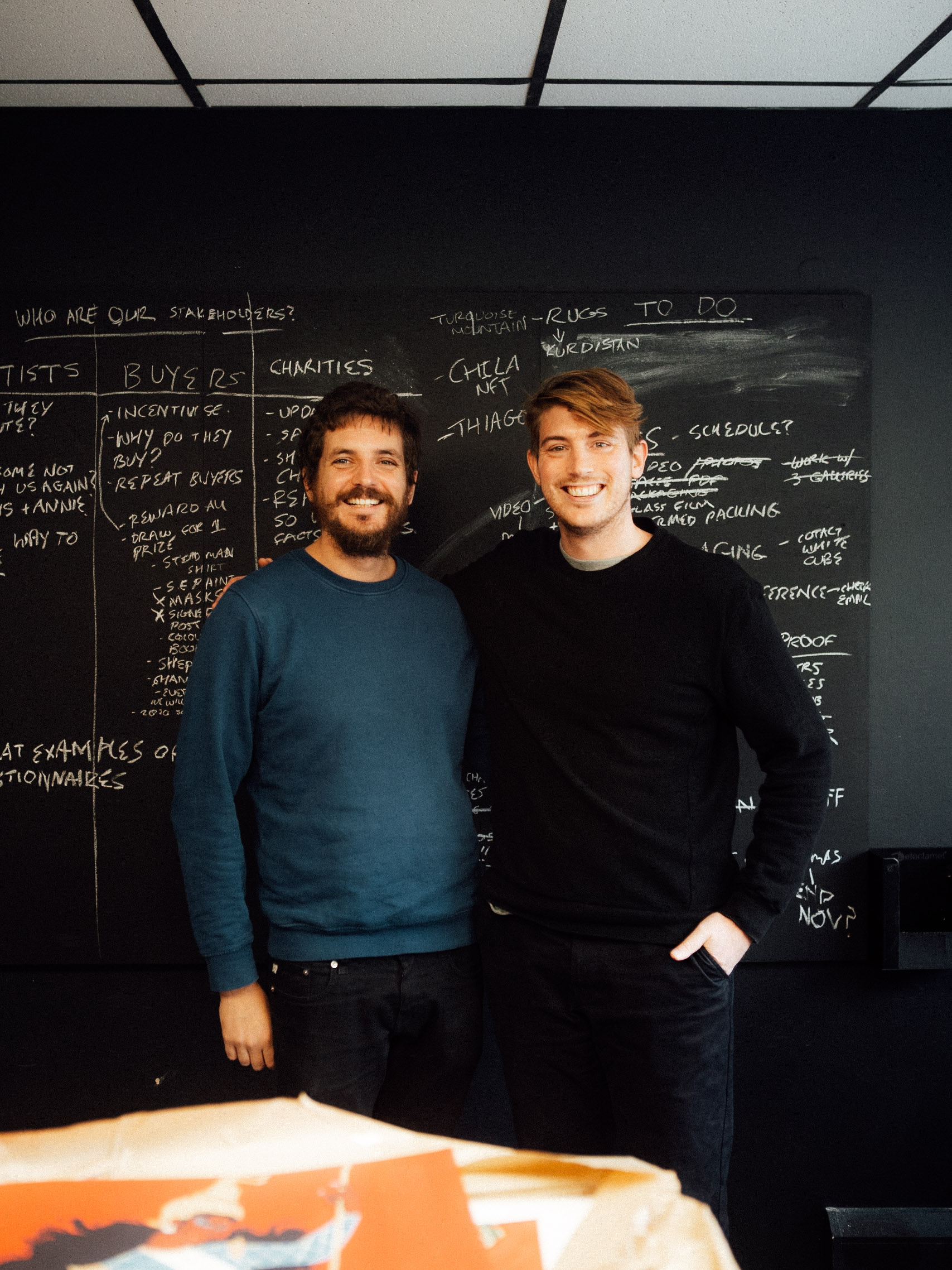 Two smiling men standing side by side in front of a blackboard filled with handwritten notes and lists.