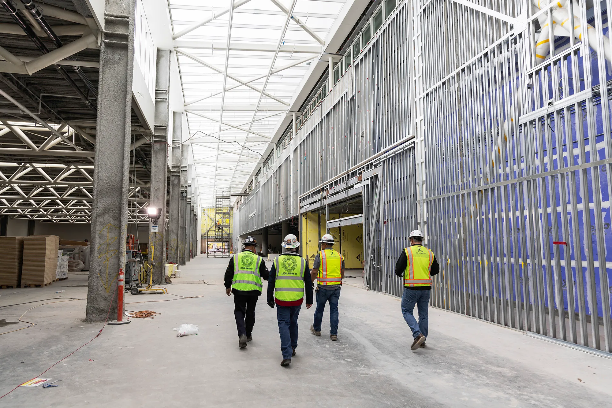 Four construction workers wearing safety vests and helmets walking inside a large, unfinished building under construction.