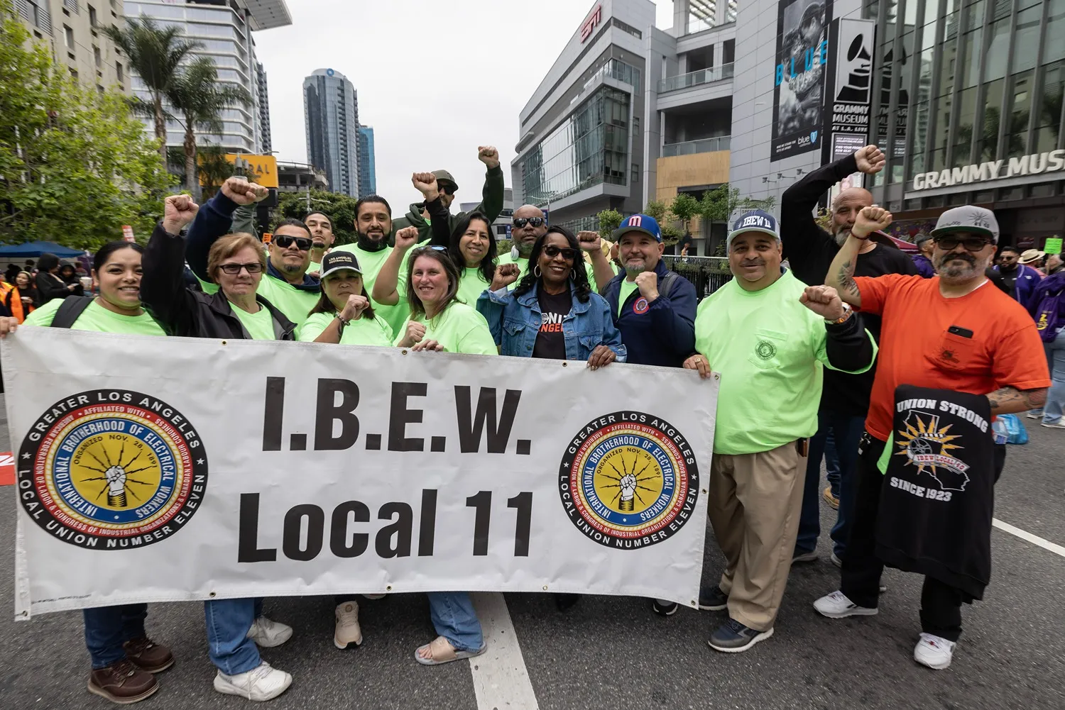 Group of union members from I.B.E.W. Local 11 holding a banner and raising their fists on a city street near the Grammy Museum.