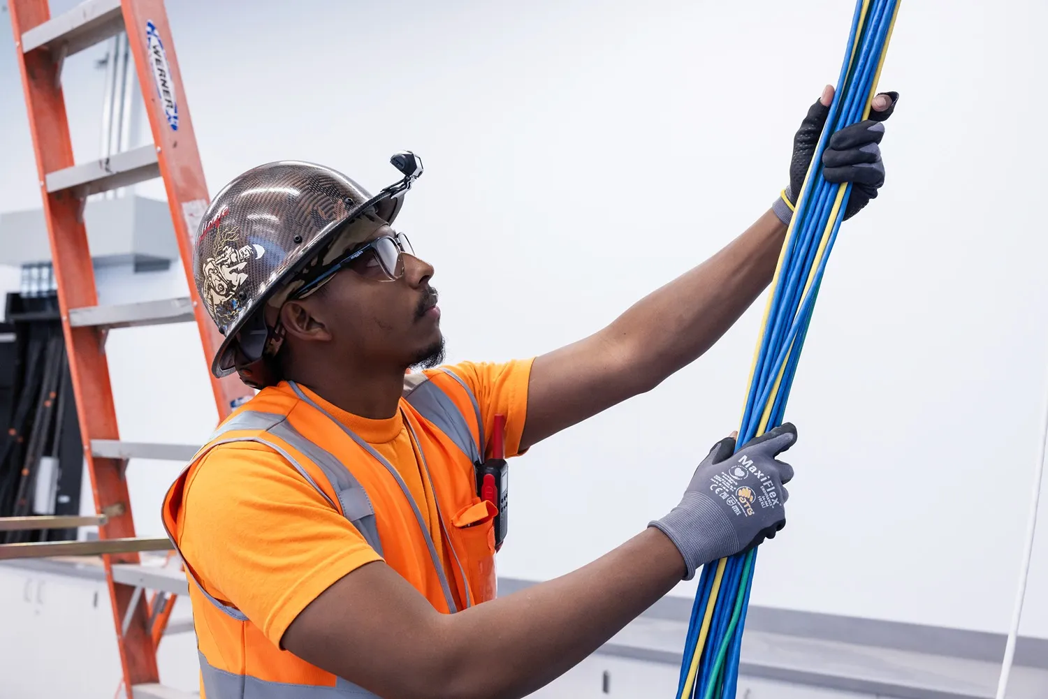 Construction worker in orange safety vest and helmet holding blue and yellow cables indoors.