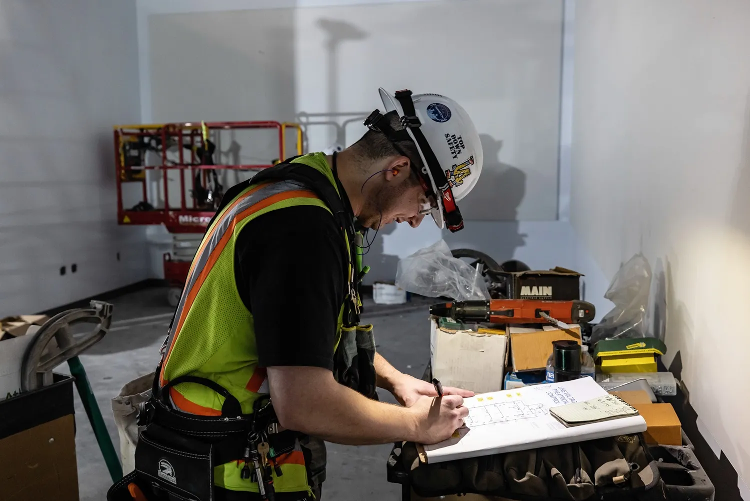 Construction worker wearing a hard hat and safety vest writing on building plans inside a construction site.