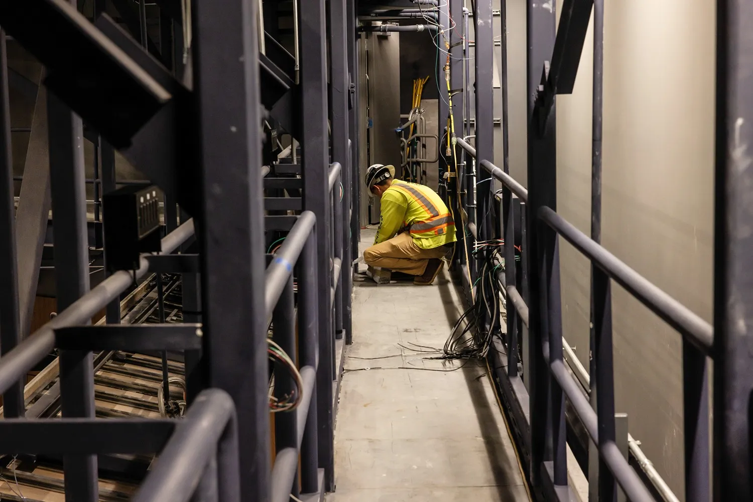 Worker in safety vest and helmet crouching on a narrow industrial walkway surrounded by metal railings and cables.