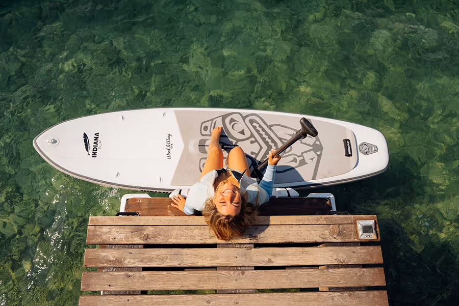 Frau auf einem SUP-Board, das neben einem Holzsteg im klaren Wasser liegt, schaut nach oben und hält eine Paddel.