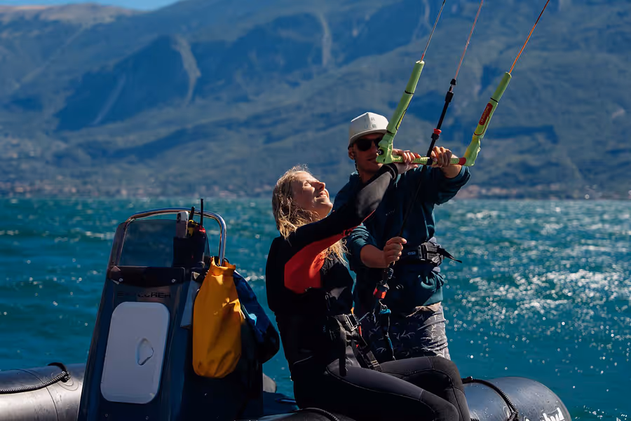Zwei Personen in Neoprenanzügen auf einem Schlauchboot, eine zeigt einer anderen den Umgang mit einer Kitesurf-Leash vor blauem Wasser und Bergen im Hintergrund.