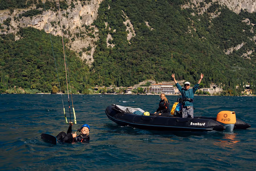 Frau beim Kitesurfen auf dem Wasser neben einem schwarzen Schlauchboot mit zwei Personen und grünen bewaldeten Bergen im Hintergrund.
