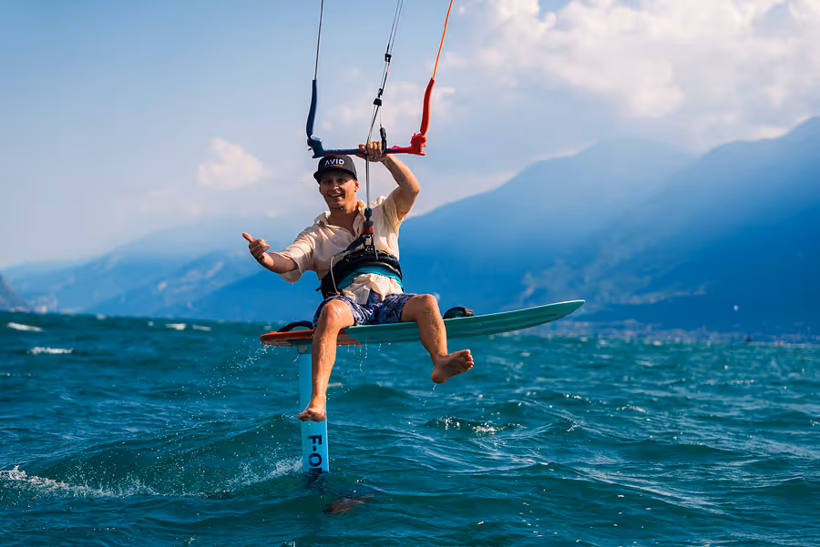 Ein Mann sitzt auf einem Foilboard und hält ein Lenkdrachen-Halterungsgriff über einem See mit Bergen im Hintergrund.
