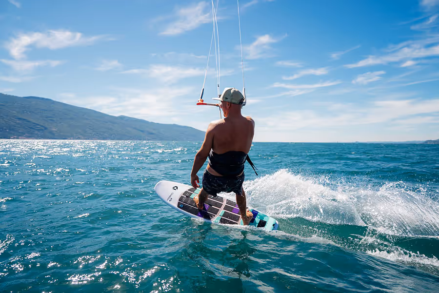 Mann, der auf einem Kiteboard über den Gardasee fährt, unter blauem Himmel mit Bergen im Hintergrund.