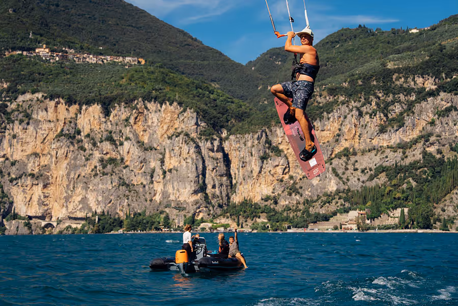 Kitesurfer springt über das blaue Wasser eines Sees, im Hintergrund Felsen und grüne Hügel.