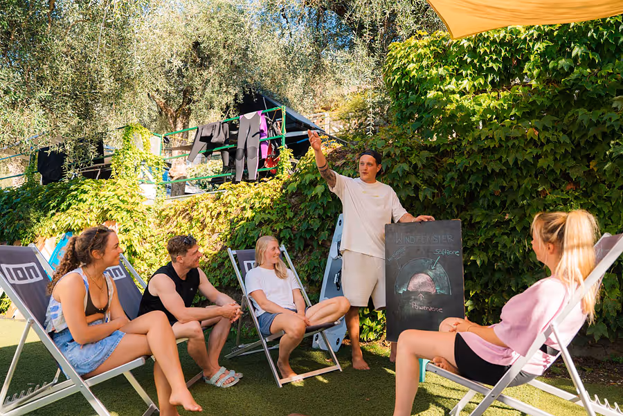Gruppe von vier Personen sitzt auf Liegestühlen im Freien, während eine fünfte Person eine Tafel mit einer Skizze hält und erklärt.