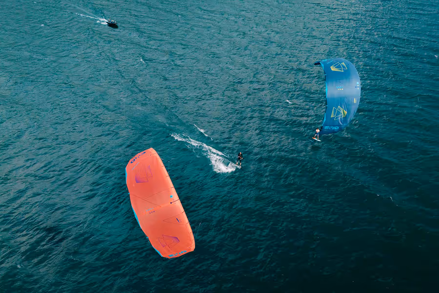Zwei Kitesurfer mit einem roten und einem blauen Kite, die auf offener See fahren, begleitet von einem kleinen Boot.