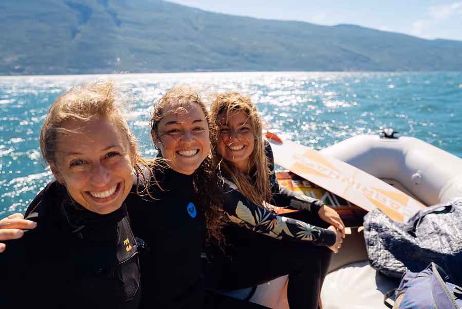 Drei lächelnde Frauen in Neoprenanzügen sitzen in einem Boot mit Berglandschaft und glitzerndem Wasser im Hintergrund.