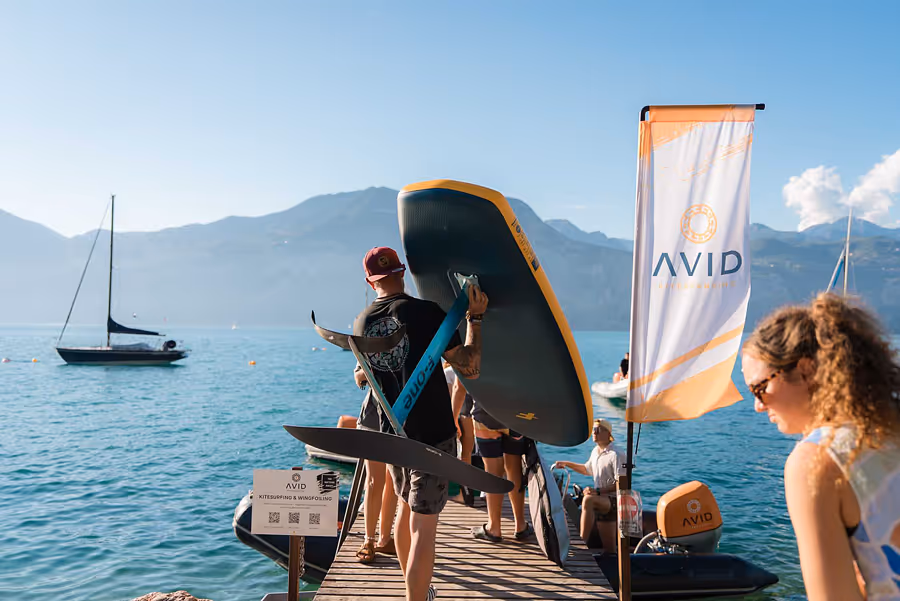 Person trägt ein Foilboard auf einem Steg am See mit Bergen im Hintergrund, daneben ein AVID-Banner.