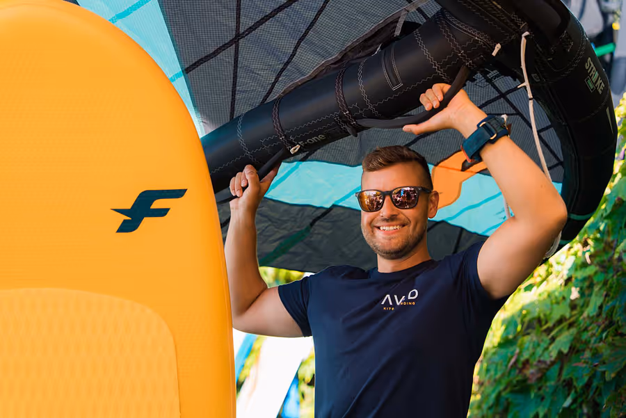 Mann mit Sonnenbrille hält ein aufblasbares Segel und steht neben einem gelben Foilbrett.