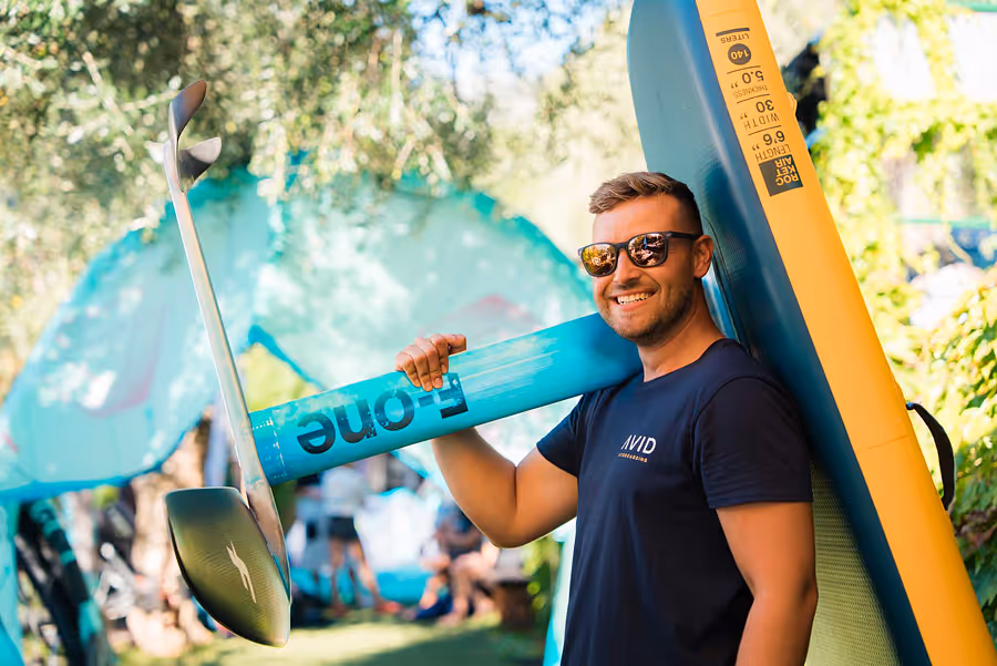 Lächelnder Mann mit Sonnenbrille trägt ein blaues F-One Foil auf der Schulter, umgeben von grünem, bewaldetem Hintergrund.