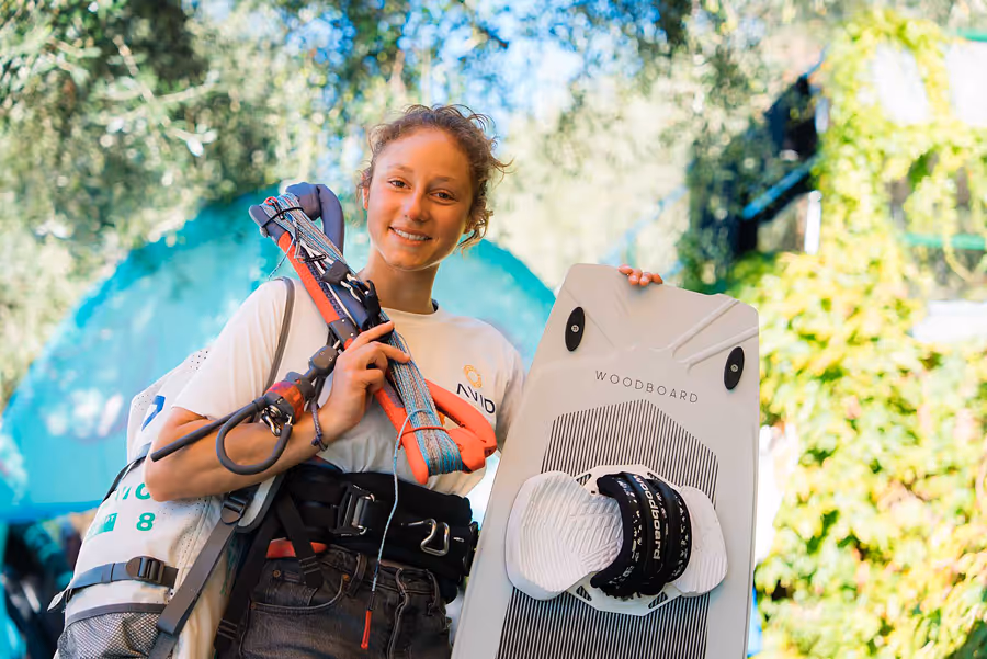Junge Frau hält ein Kiteboard und Trapezseile, lächelnd vor grünem, verschwommenem Hintergrund.