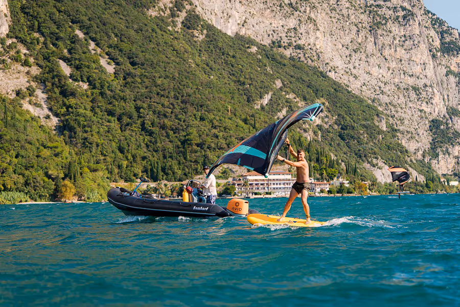 Mann auf einem gelben Foilboard mit Wing-Segel, begleitet von einem Schlauchboot auf einem See vor bergiger, grüner Landschaft.
