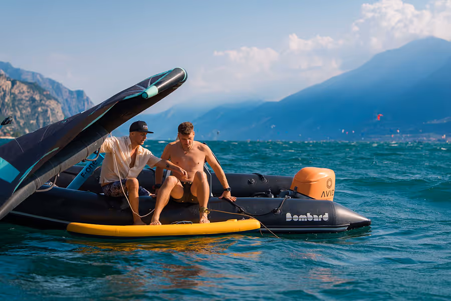 Zwei Männer auf einem schwarzen Schlauchboot auf dem Wasser, umgeben von Bergen und blauem Himmel.