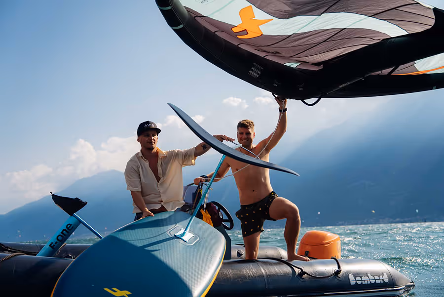 Zwei Männer auf einem Schlauchboot auf einem See, einer hält ein großes Trapez-Segel, der andere sitzt neben einem Hydrofoil-Surfbrett.