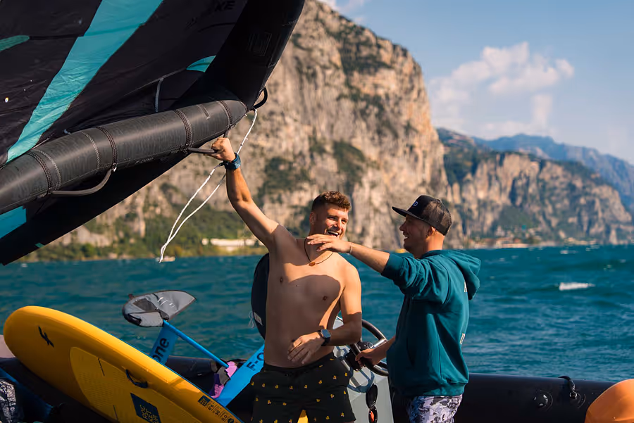 Zwei junge Männer auf Schlauchboot mit Bergkulisse, einer hält ein Wing und lächelt.