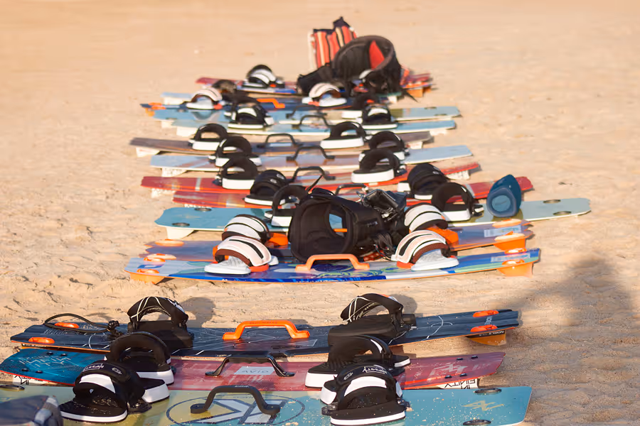 Mehrere Kitesurf-Boards mit Fußschlaufen liegen nebeneinander auf sandigem Boden.