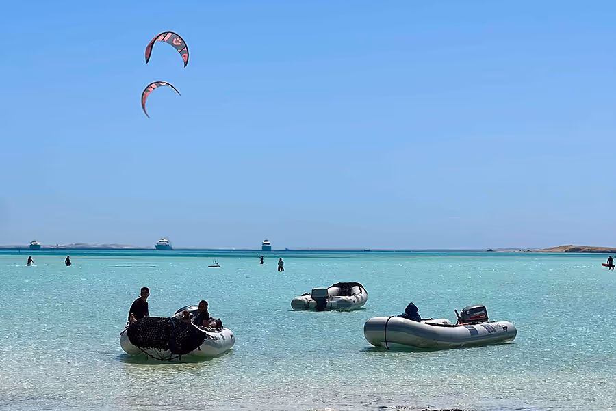 Leute auf kleinen Schlauchbooten im klaren türkisfarbenen Wasser unter blauem Himmel mit zwei roten Kites am Himmel.