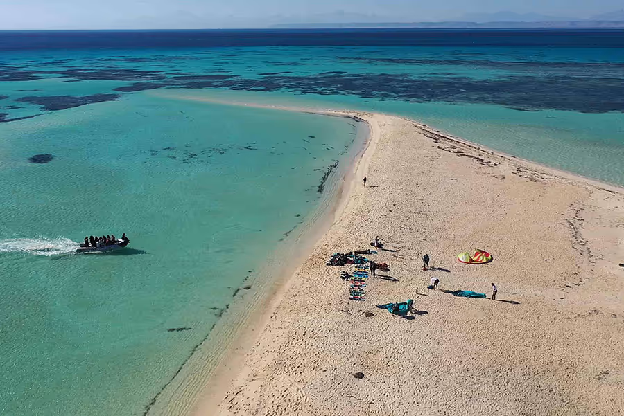 Drohnenaufnahme einer sandigen Landzunge, umgeben von klarem türkisfarbenem Wasser, mit einer kleinen Gruppe von Menschen und einem Boot in der Nähe.