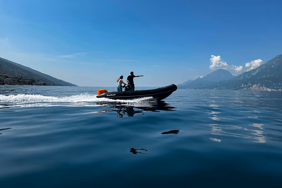 Zwei Personen fahren in einem Schlauchboot über ruhigen See vor bergiger Landschaft bei klarem Himmel.