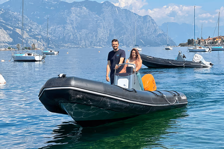 Zwei Personen auf einem schwarzen Schlauchboot in einem See mit Bergen und Segelbooten im Hintergrund.