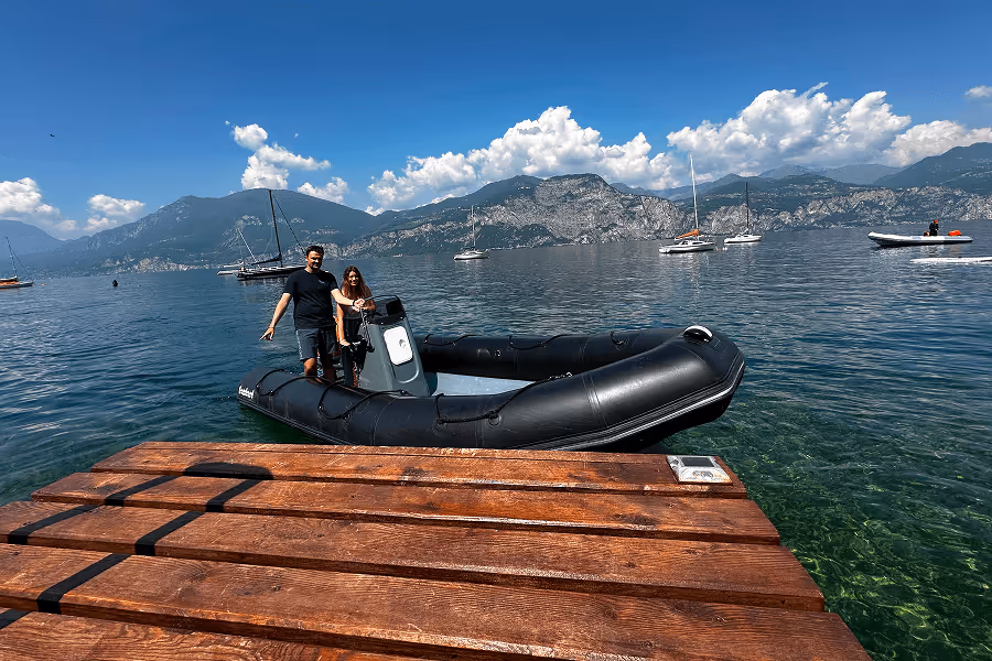 Zwei Personen stehen in einem schwarzen Schlauchboot am Holzsteg auf klarem Wasser mit Bergen im Hintergrund.