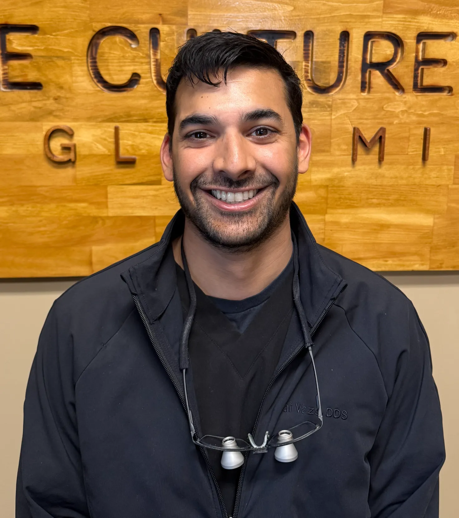 Smiling man wearing a dark jacket and dental loupes around his neck, standing in front of a wooden sign.