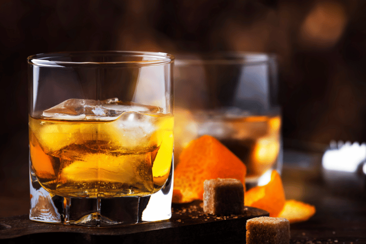 Close-up of a whiskey glass with ice, orange peel, and sugar cubes on a dark background.