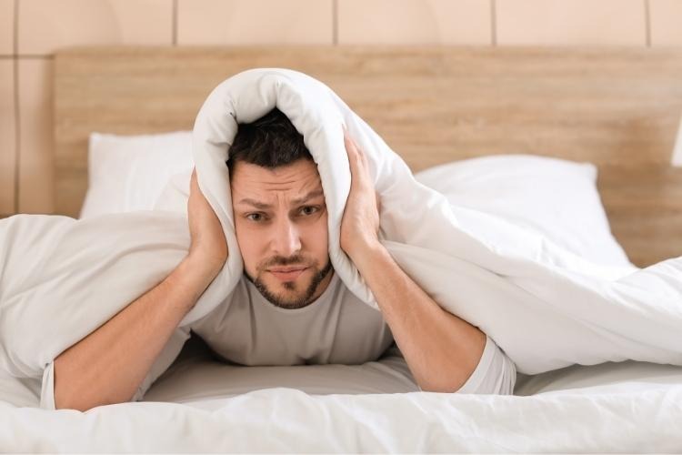 Man with his head under the duvet looking unhappy.