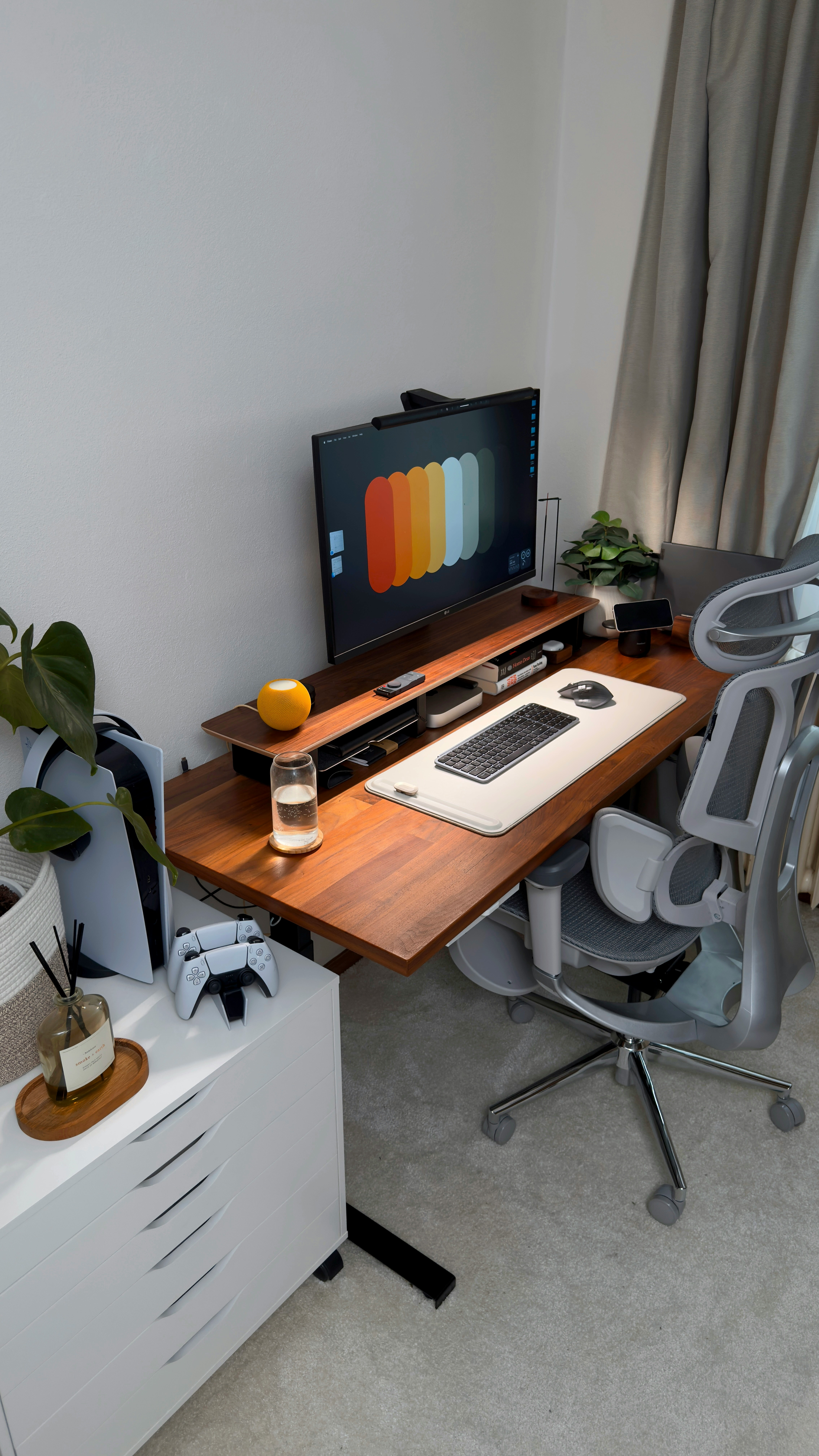Espacio de trabajo limpio con escritorio de madera, monitor con gráficos coloridos, teclado inalámbrico, silla ergonómica y consola de videojuegos en una cómoda blanca.