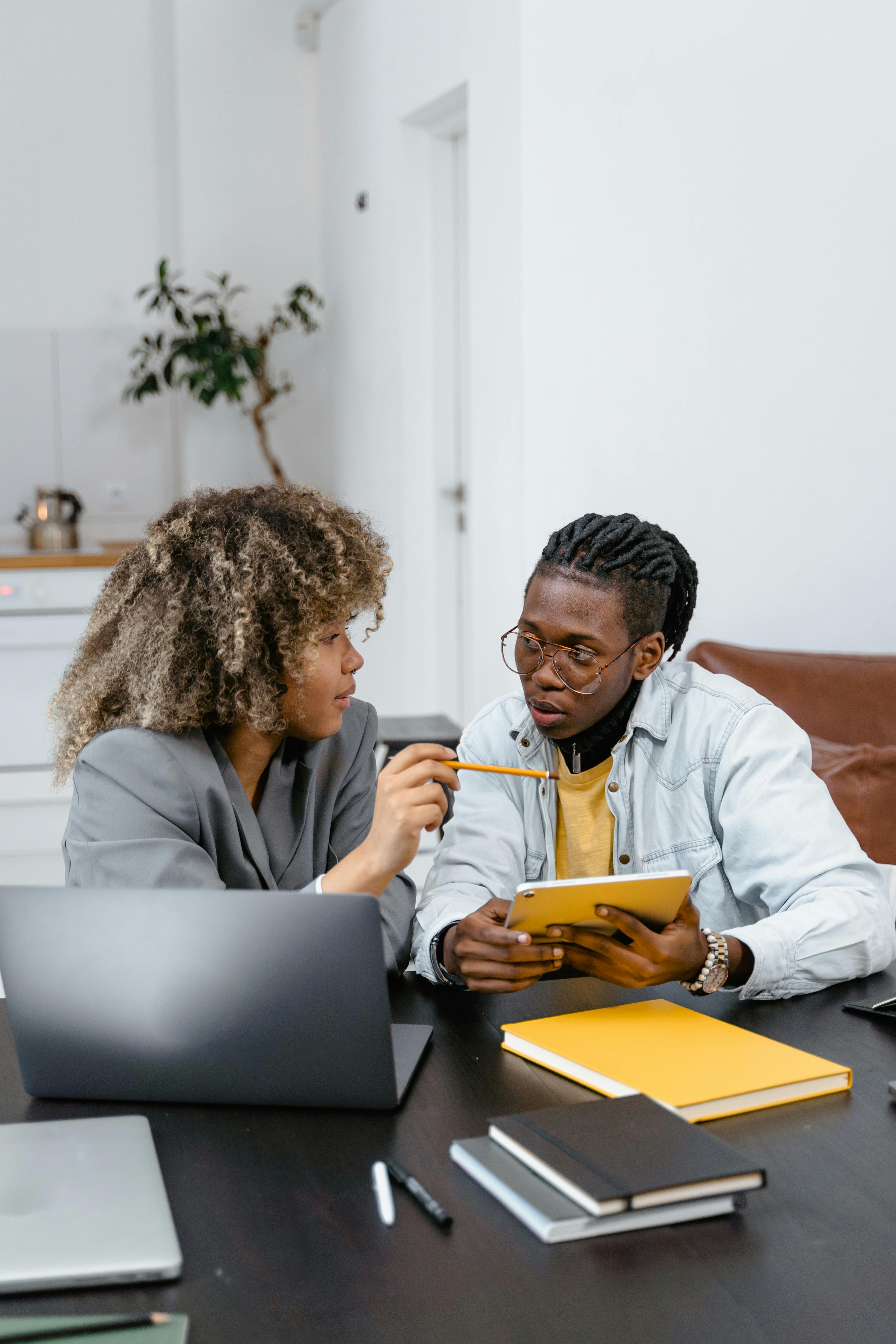 Dos jóvenes discutiendo mientras trabajan con una tableta y un portátil en una mesa con libros y bolígrafos.