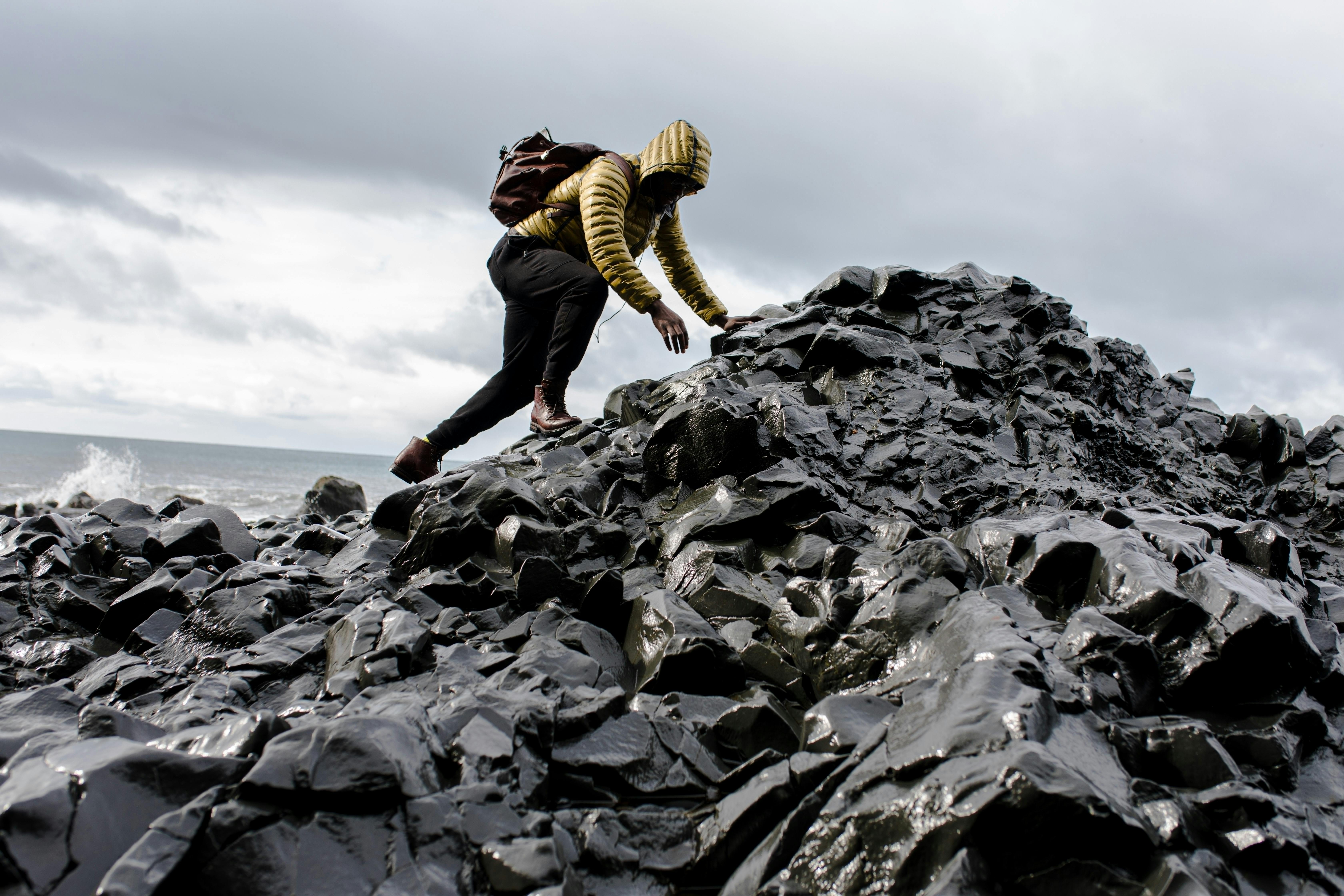 Persona con chaqueta amarilla y mochila subiendo rocas negras cerca del mar bajo un cielo nublado.