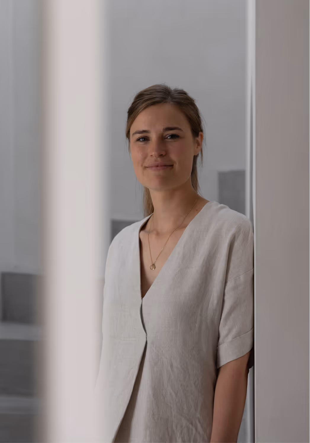 Vrouw met lichtbruine haren en een beige blouse die zacht glimlacht in een minimalistische kamer.