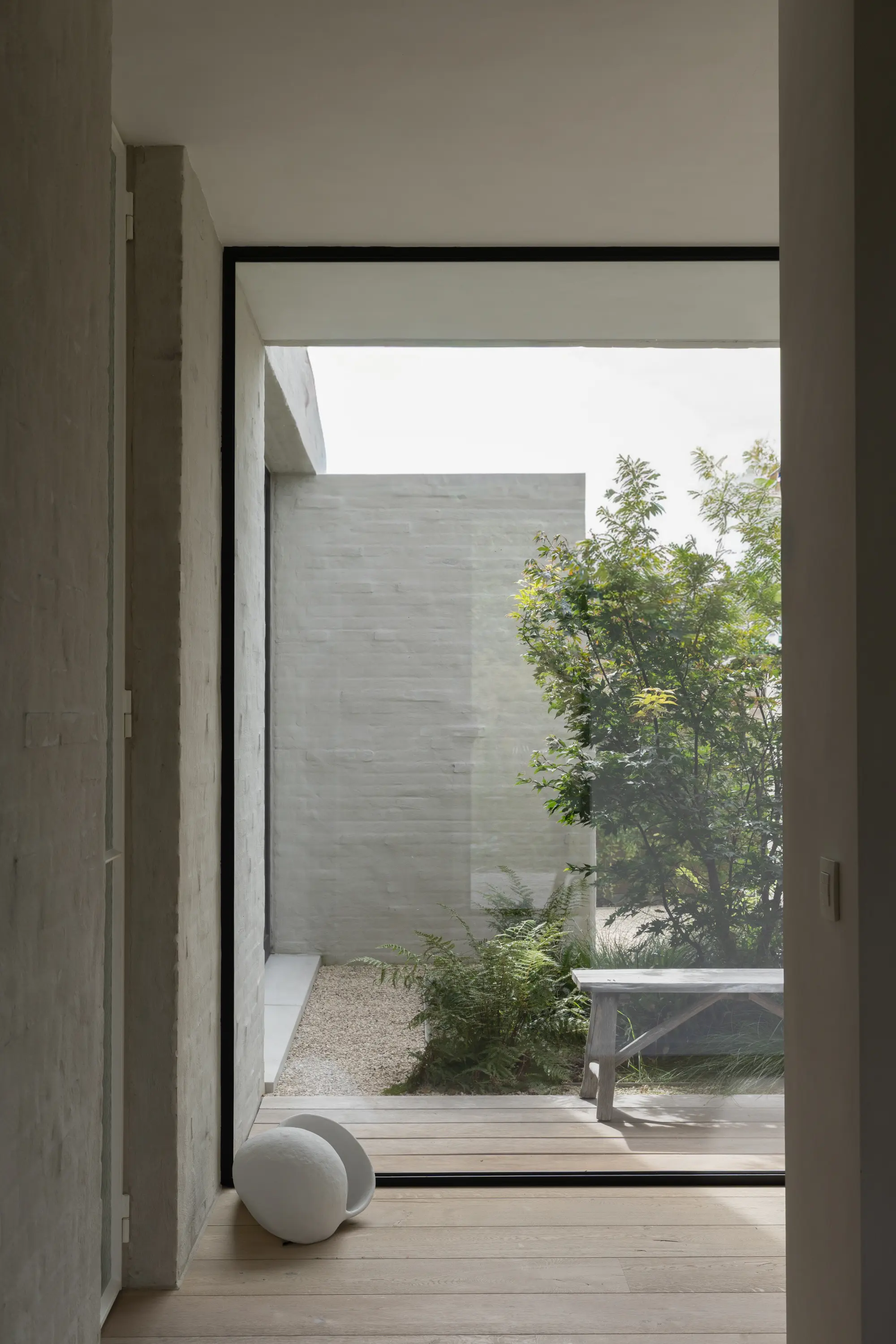 Modern interieur met groot raam dat uitkijkt op een kleine tuin met varens, een houten bank en een witte muur.