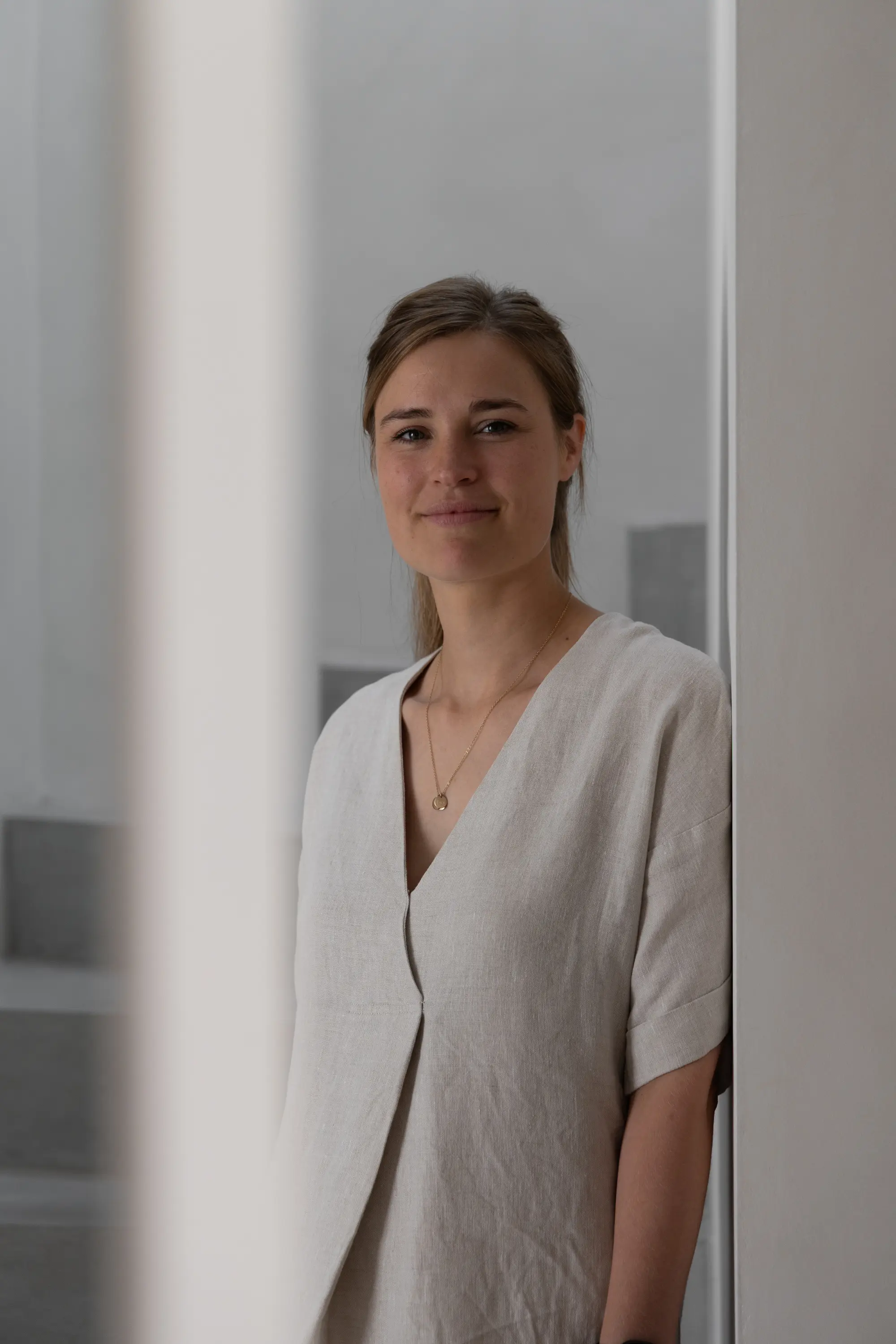 Architect Laura Engelen met lichtbeige tuniek en gouden ketting die tegen een witte muur staat en glimlacht.
