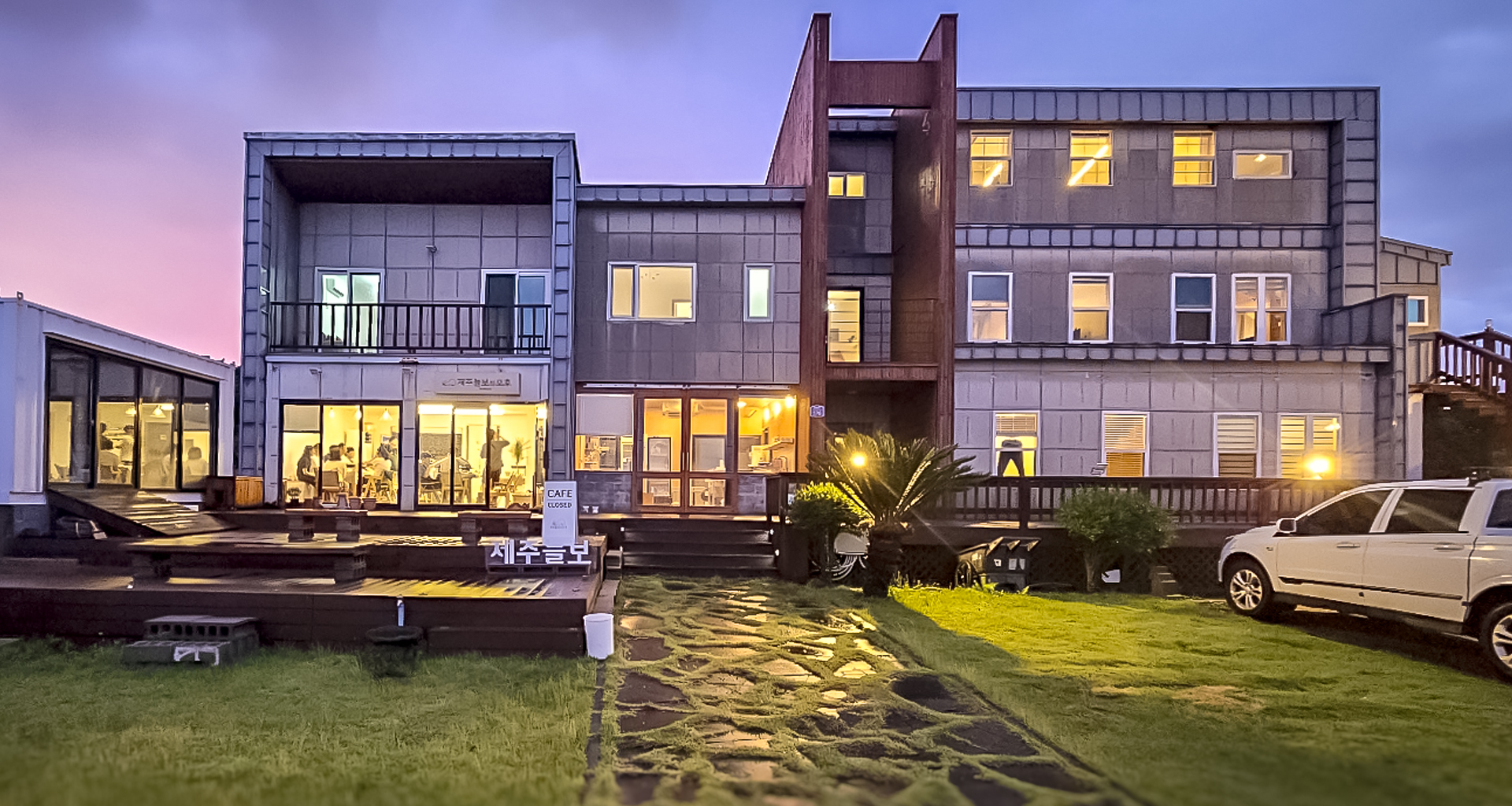 '제주늘보의오후' 저녁 시간 시설 정면에서 본 장면으로, 시설이 오렌지색 조명에 둘러쌓여있다. This is a scene seen from the front of the facility in the evening of 'JEJUNEULBO' and the facility is surrounded by orange lights.