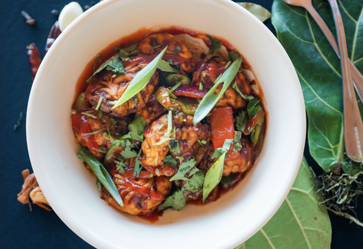 A bowl of spicy Indo-Chinese chicken manchurian garnished with green onions and cilantro, placed on a dark surface with leafy greens and cutlery nearby.