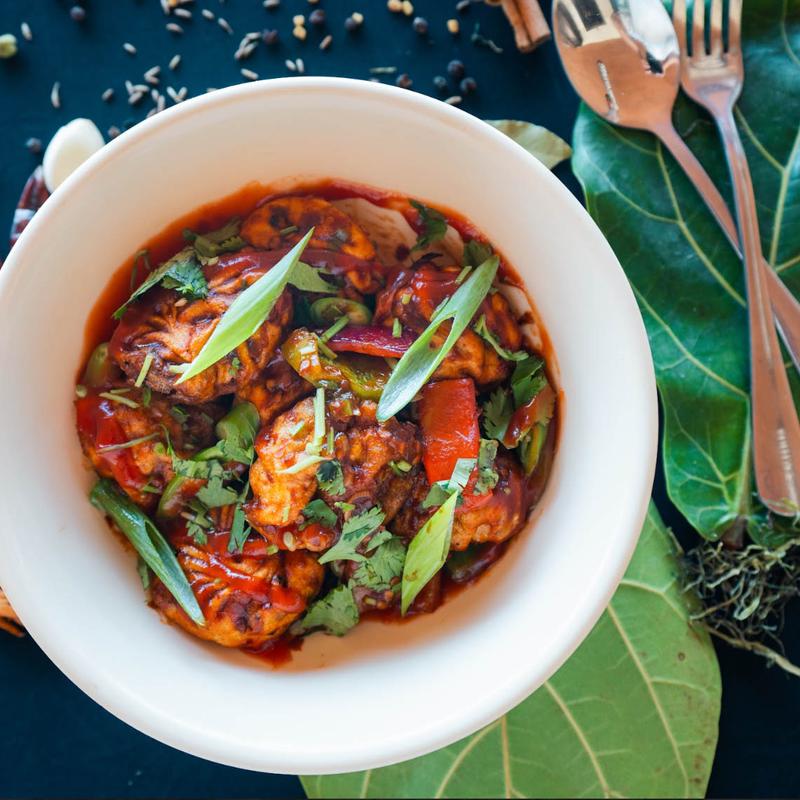 A white bowl filled with spicy Manchurian, garnished with spring onions and cilantro. Surrounded by green leaves and cutlery, creating a fresh, flavor-rich scene.