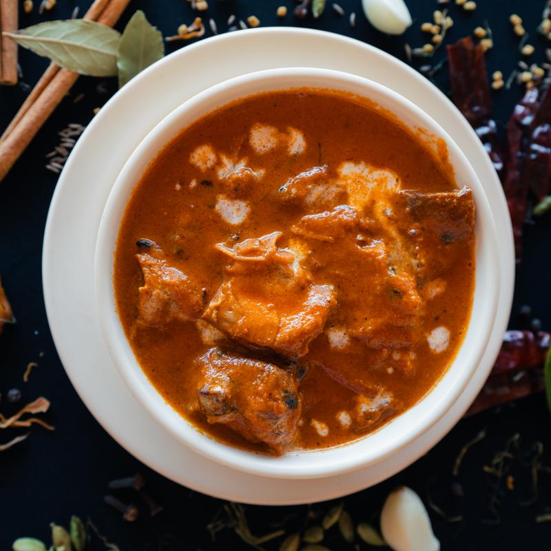 A bowl of rich, red curry with tender meat chunks, garnished with cream swirls. Set against a black background with spices, evoking warmth and flavor.