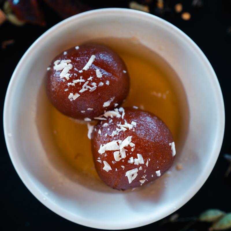 Two round gulab jamuns in syrup sit in a white bowl, sprinkled with coconut flakes. The rich brown color and syrupy texture evoke indulgence.